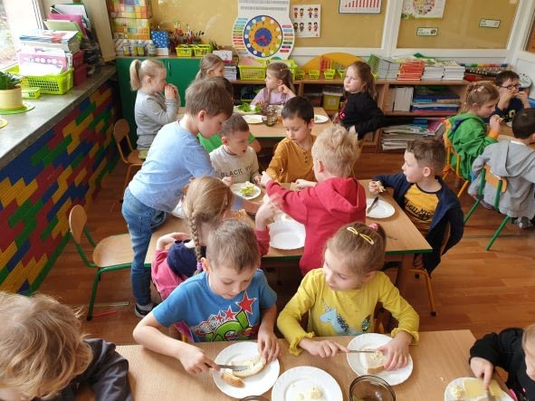 Dzieci siedzą przy stolikach, smarują kromki chleba masłem.